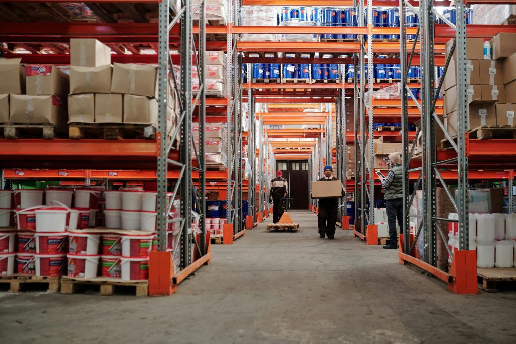 Armazém com trabalhadores movimentando caixas e insumos de obra comercial em prateleiras industriais.
