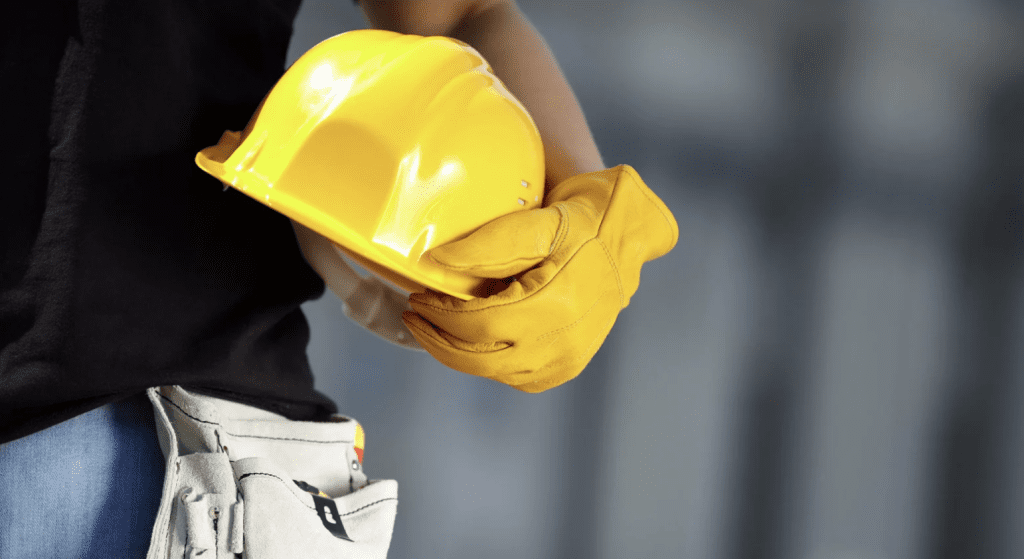Trabalhador da construção civil de uniforme preto e jeans segurando um capacete amarelo com luvas de proteção, simbolizando segurança no canteiro de obras.