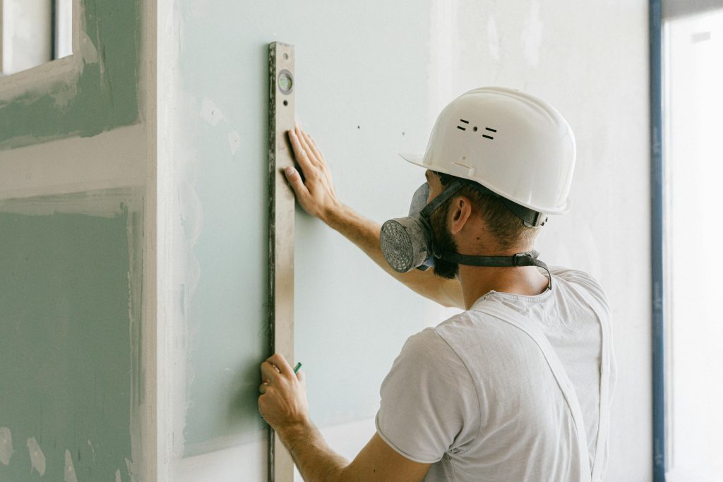 Engenheiro usando capacete e máscara mede o alinhamento de uma parede, simbolizando a responsabilidade técnica exigida em obras de franquias.