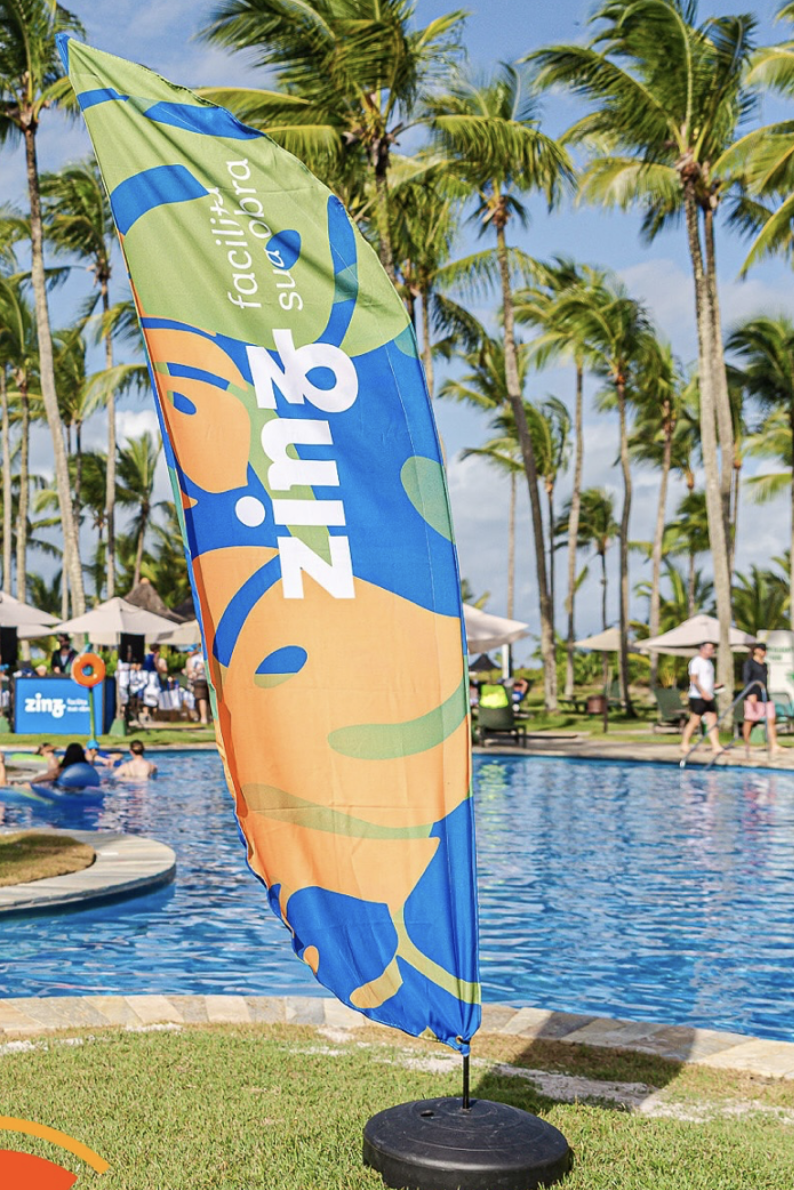 Bandeira da Zinz com as cores verde, azul e laranja posicionada à beira da piscina durante a Convenção ABF 2025 em Comandatuba, com palmeiras e congressistas ao fundo.